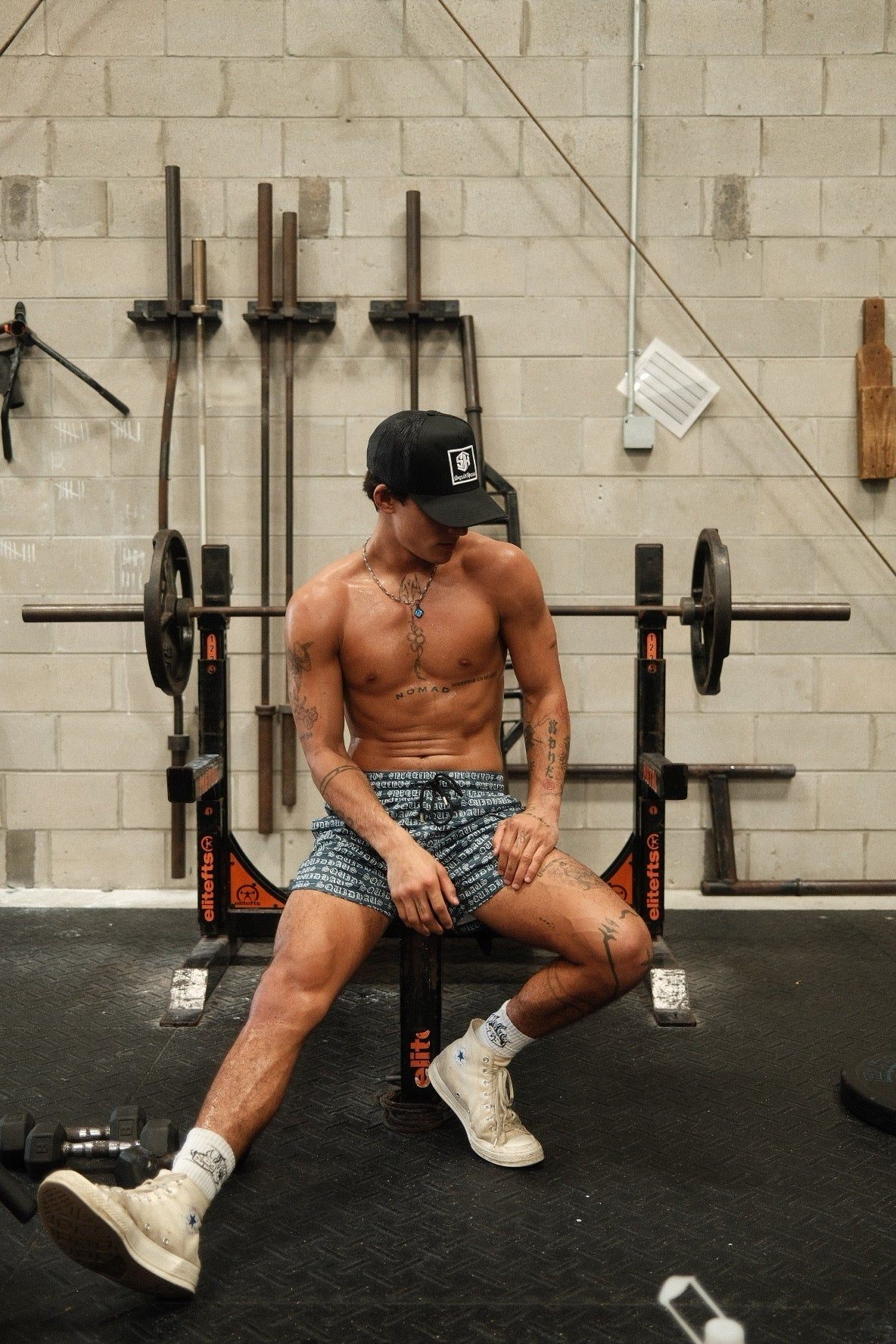 A bodybuilder at the gym sitting on the benchpress machine wearing 4 inch inseam mesh shorts and a trucker hat in black, both by SquidHaus