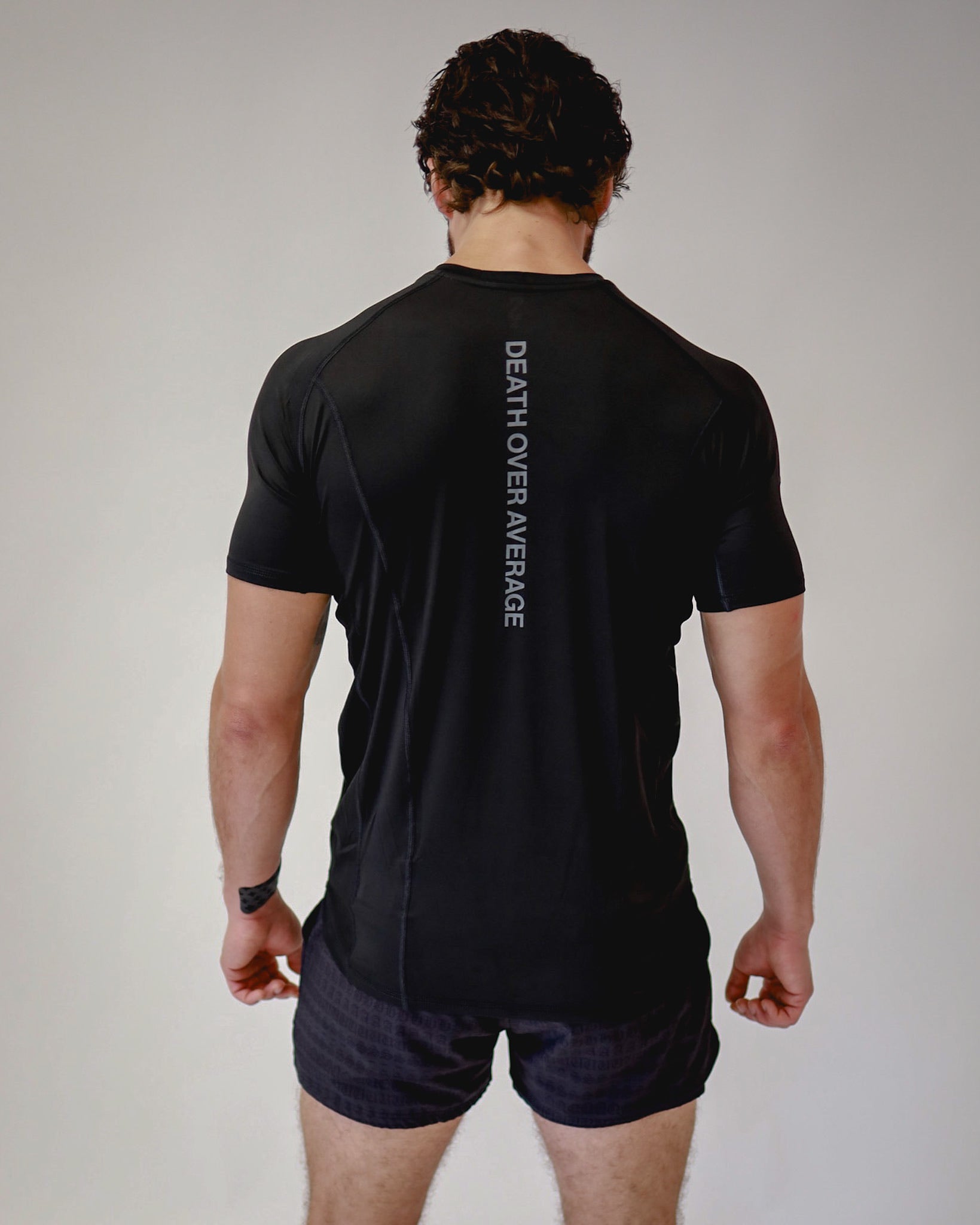 Rear view of an athlete wearing a Pro-Tech V1 athletic fitted short sleeve t-shirt in black  showing the "Death Over Average" slogan on the back, with 4" inseam mesh shorts, from SquidHaus