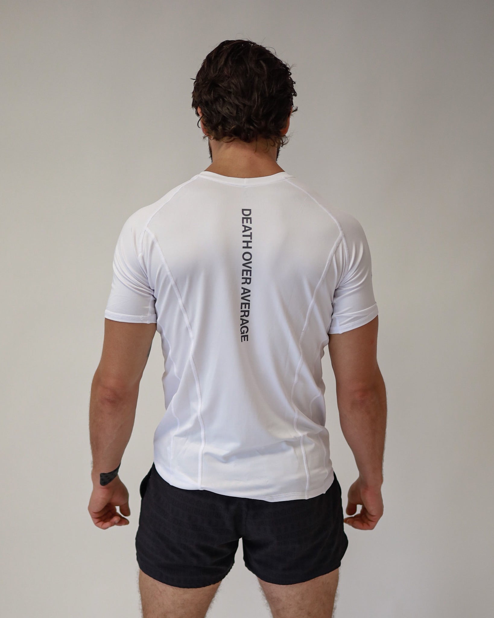 Rear view of an athlete wearing a Pro-Tech V1 athletic fitted short sleeve t-shirt in white showing the "Death Above Average" slogan on the back, with 4" inseam mesh shorts in black, from SquidHaus