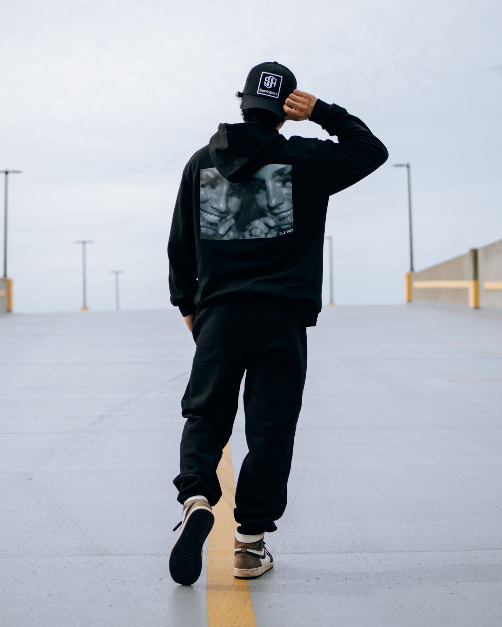 A man walking away from the camera while wearing SquidHaus' Death Over Average sweatpants, Lip Tat Hoodie and black trucker hat.