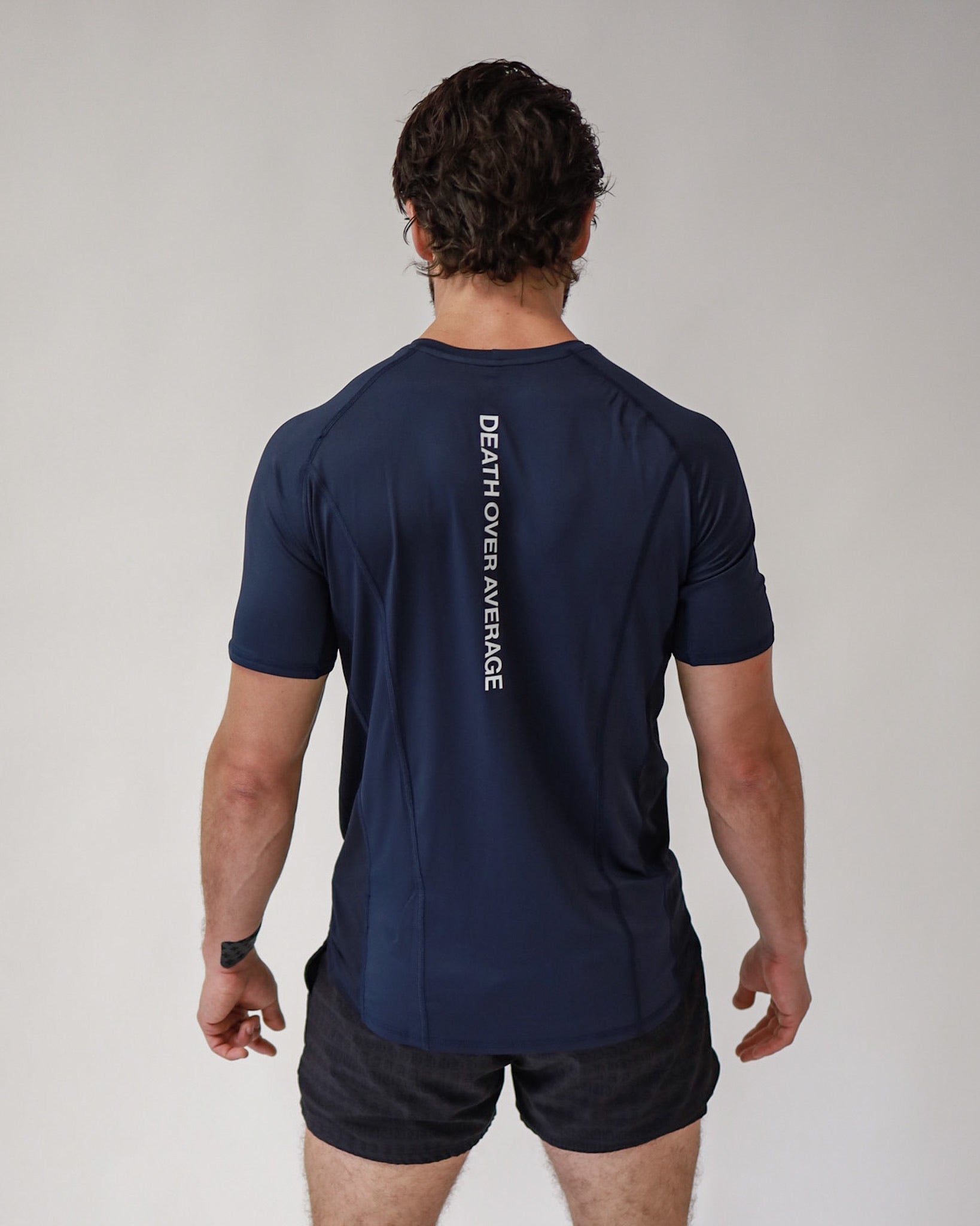 Rear view of an athlete wearing a Pro-Tech V1 athletic fitted short sleeve t-shirt in navy blue showing the "Death Above Average" slogan, with 4" inseam mesh shorts in black, from SquidHaus