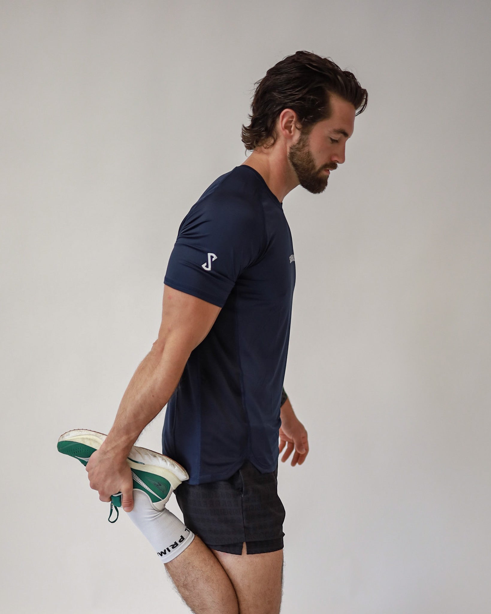 A side view of a runner stretching before his run while wearing a Pro-tech V1 athletic fitted short sleeve t-shirt in navy blue with 4" inseam mesh shorts in black, from SquidHaus