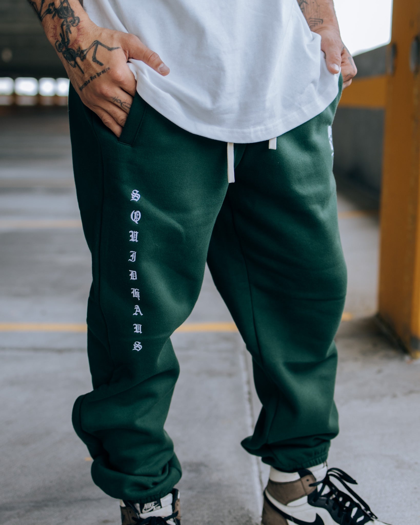 A man wearing SquidHaus Death Over Average sweatpants in green, showing the "SquidHaus" embroidery on the right pant leg