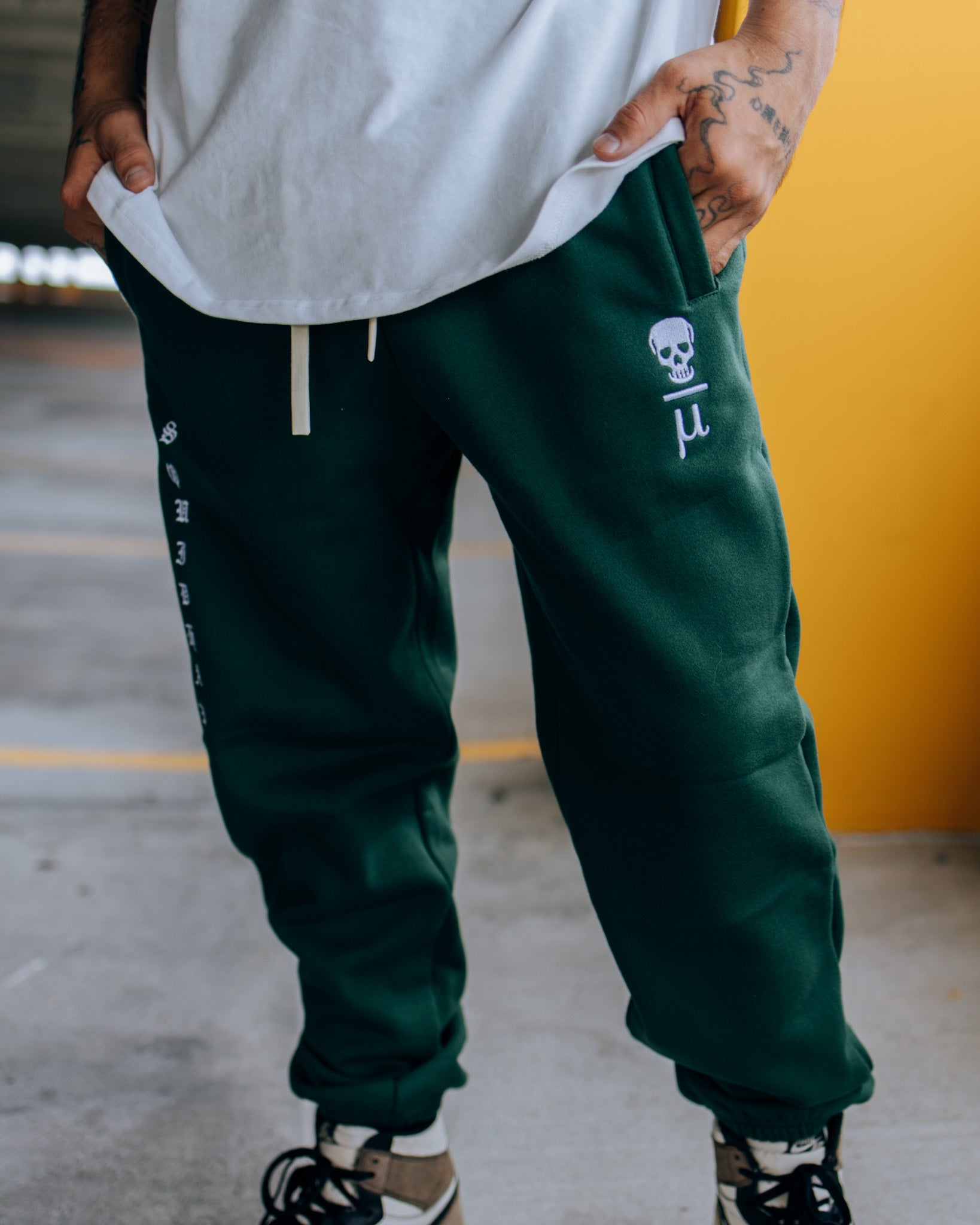 A man wearing SquidHaus Death Over Average sweatpants in green, showing the Death Over Average logo on the top of the left pant leg