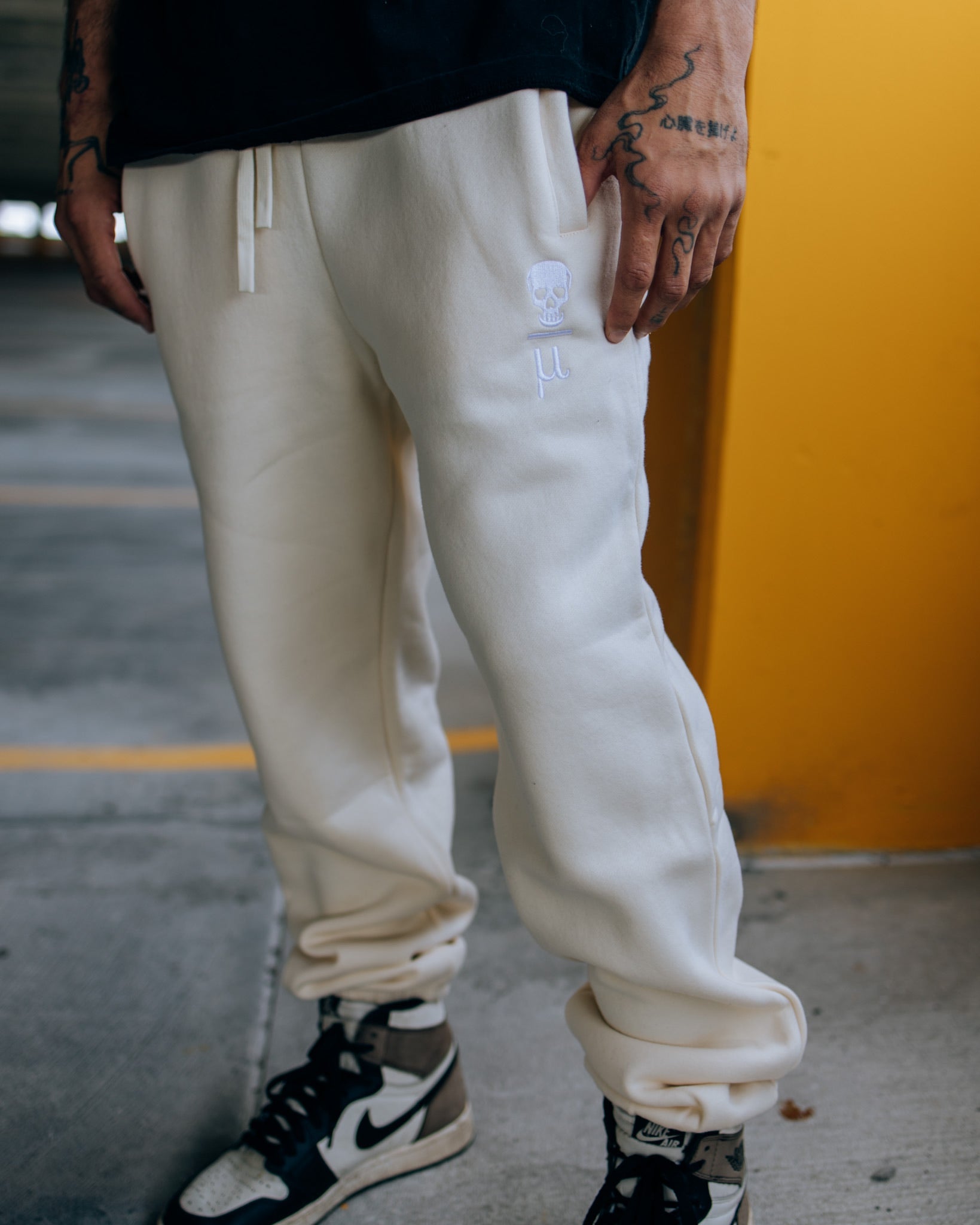 A man wearing SquidHaus Death Over Average sweatpants in white, showing the Death Over Average logo on the top of the left pant leg